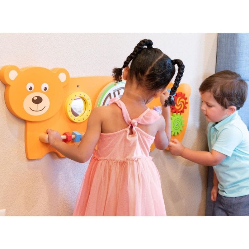 A young girl in a pink dress and a young boy in a light blue shirt play with a colorful bear-shaped wall activity board on a beige wall.