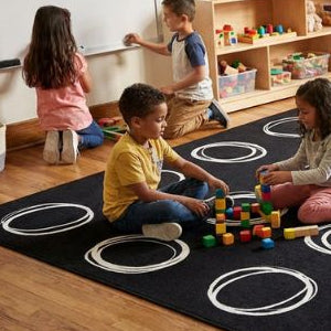 Children sitting on a black rug with white circles in a classroom setting.