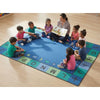 A large blue children's rug with colorful alphabet and shape borders is spread on a light wood floor, with an adult reading to a circle of children.