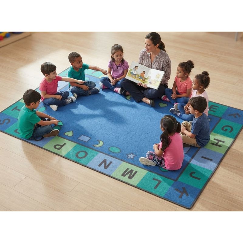 A large blue children's rug with colorful alphabet and shape borders is spread on a light wood floor, with an adult reading to a circle of children.
