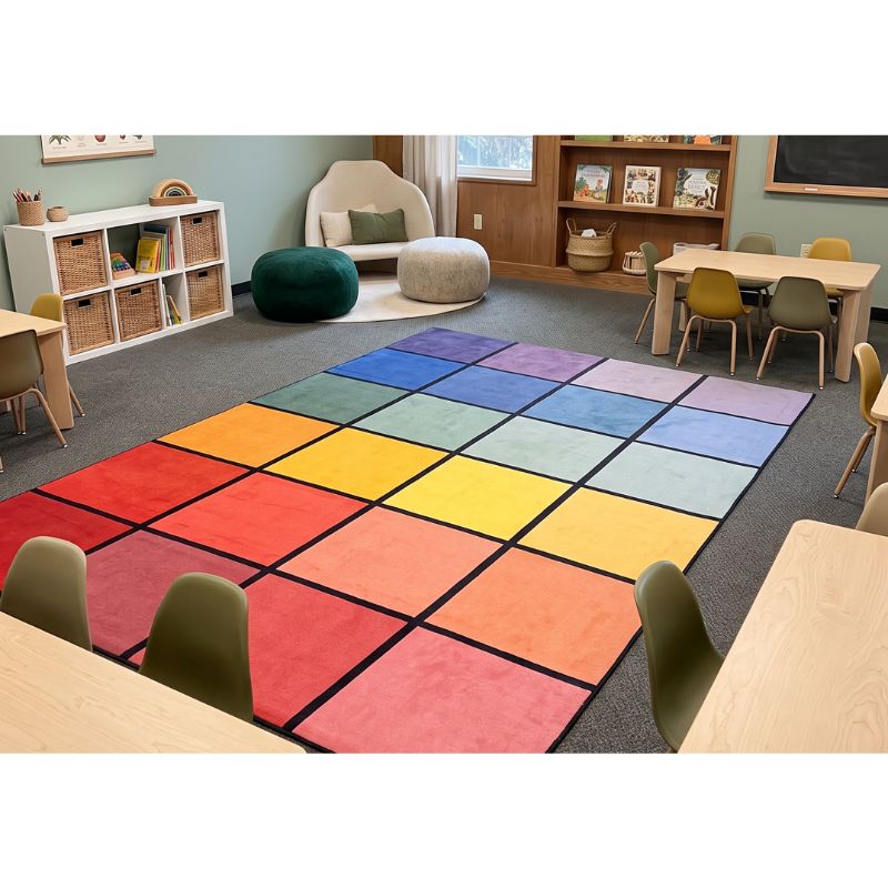A large rectangular classroom rug with a rainbow grid pattern of colored squares, shown in a modern classroom setting with tables, chairs, and storage shelves.