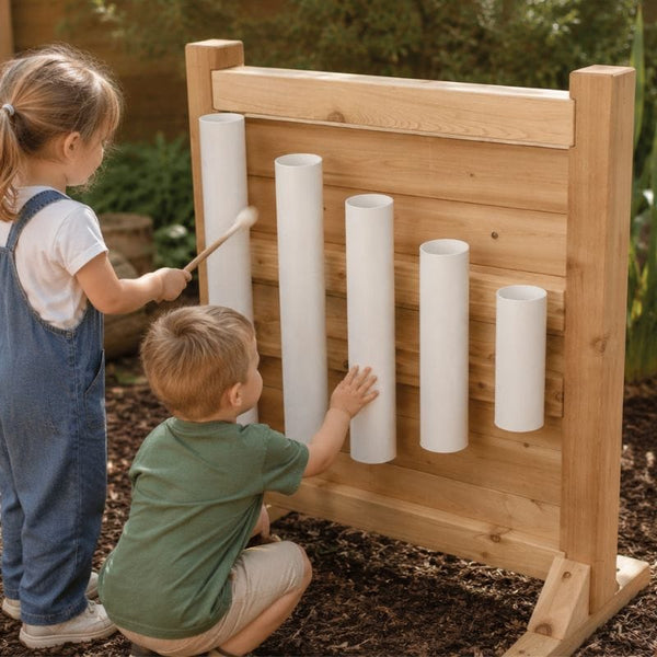 A children's outdoor water pipe toy with five vertical white PVC tubes of decreasing size mounted on a natural wood frame. A young girl in blue overalls holds a wooden mallet above the tallest tube, while a young boy in a green shirt kneels down to touch the middle tube.