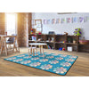A rectangular area rug with a bright blue background and a repeating pattern of smiling sunflower faces, shown in a styled children's room with wooden crates, toys, and a brick column.