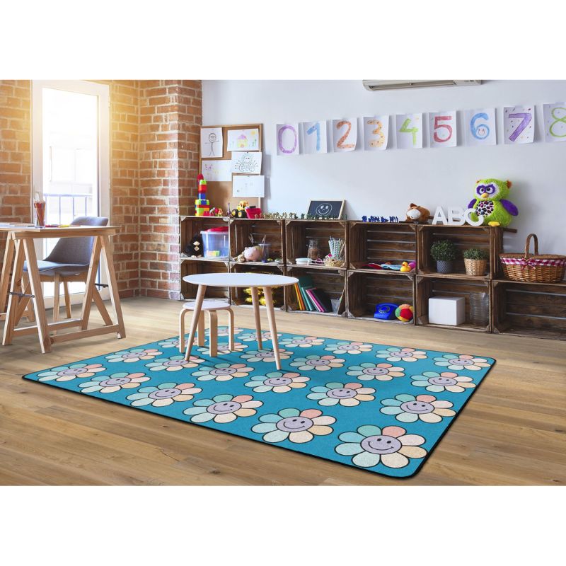 A rectangular area rug with a bright blue background and a repeating pattern of smiling sunflower faces, shown in a styled children's room with wooden crates, toys, and a brick column.
