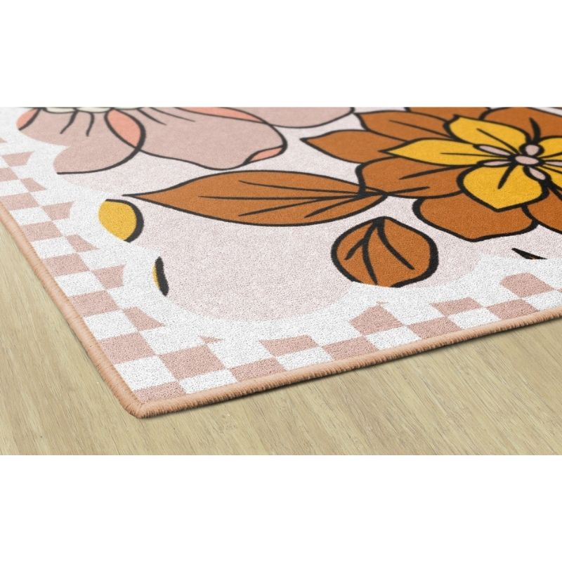 A close-up of a rectangular area rug with a floral pattern, shown on a light wood floor. The rug features a white base with large flowers in orange, yellow, and pink, brown leaves, and a pink and white checkered border.