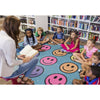 A woman sits on a small stool reading a book to a group of children sitting on a carpet with large smiley faces, in a library or bookroom with bookshelves in the background.