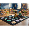 A children's classroom with kids sitting on a large black space-themed area rug decorated with colorful planets, rings, stars, and rockets. The room has blue walls, educational posters, shelves, and colorful chairs and tables.