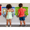 Two children standing with their backs to the camera, playing with colorful wall-mounted train-shaped activity boards on a light gray wall.