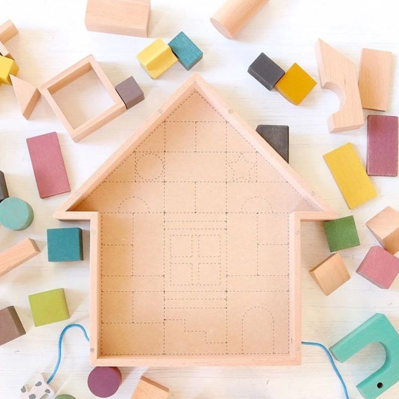 A wooden house-shaped toy with dotted maze-like line patterns on the inside, surrounded by scattered pastel wooden blocks on a light surface.