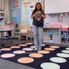 Person standing on a colorful classroom rug with educational posters and furniture in the background