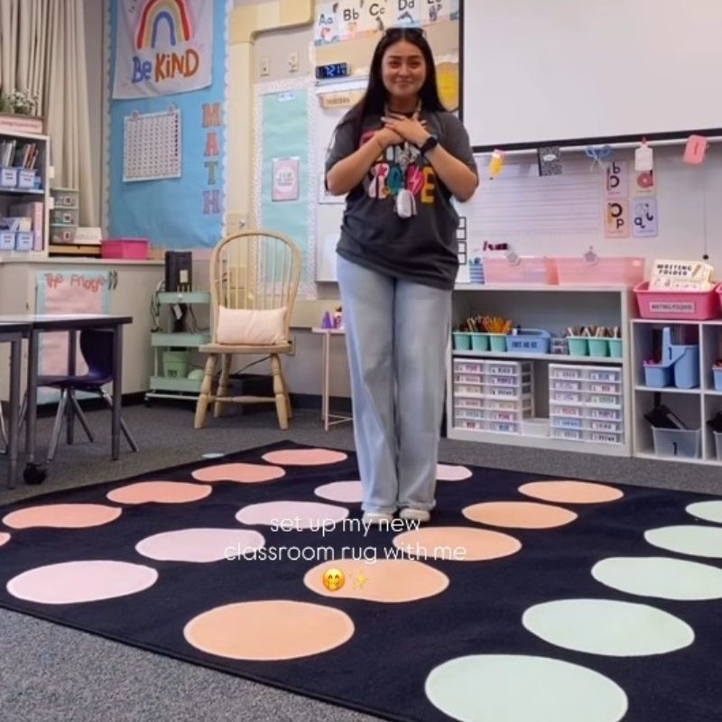 Person standing on a colorful classroom rug with educational posters and furniture in the background