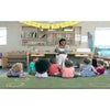 A woman sits on a desk in a children's classroom, reading to a group of young children who are sitting on a green patterned rug. The room has light wood shelves, colorful decorations, and toys.
