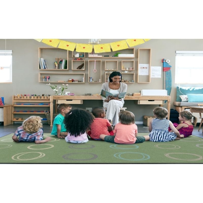 A woman sits on a desk in a children's classroom, reading to a group of young children who are sitting on a green patterned rug. The room has light wood shelves, colorful decorations, and toys.