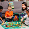 Children and a woman playing with a dinosaur toy set on a living room floor.