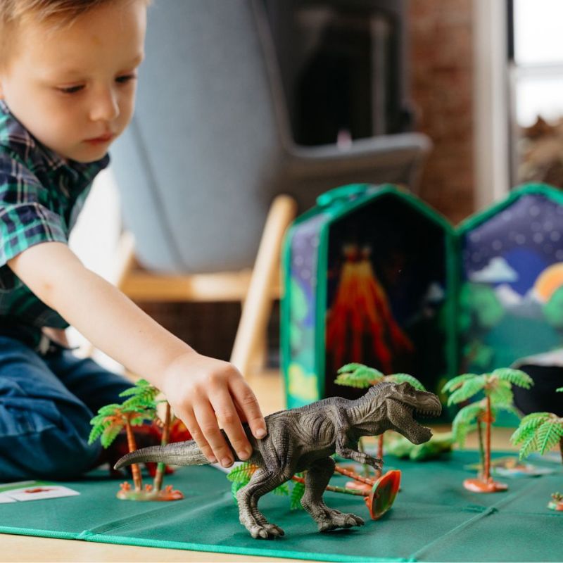 Child playing with toy dinosaur on a play mat with toy trees and volcano in the background.