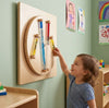 Child interacting with a wooden educational toy on a light green wall with children's drawings.